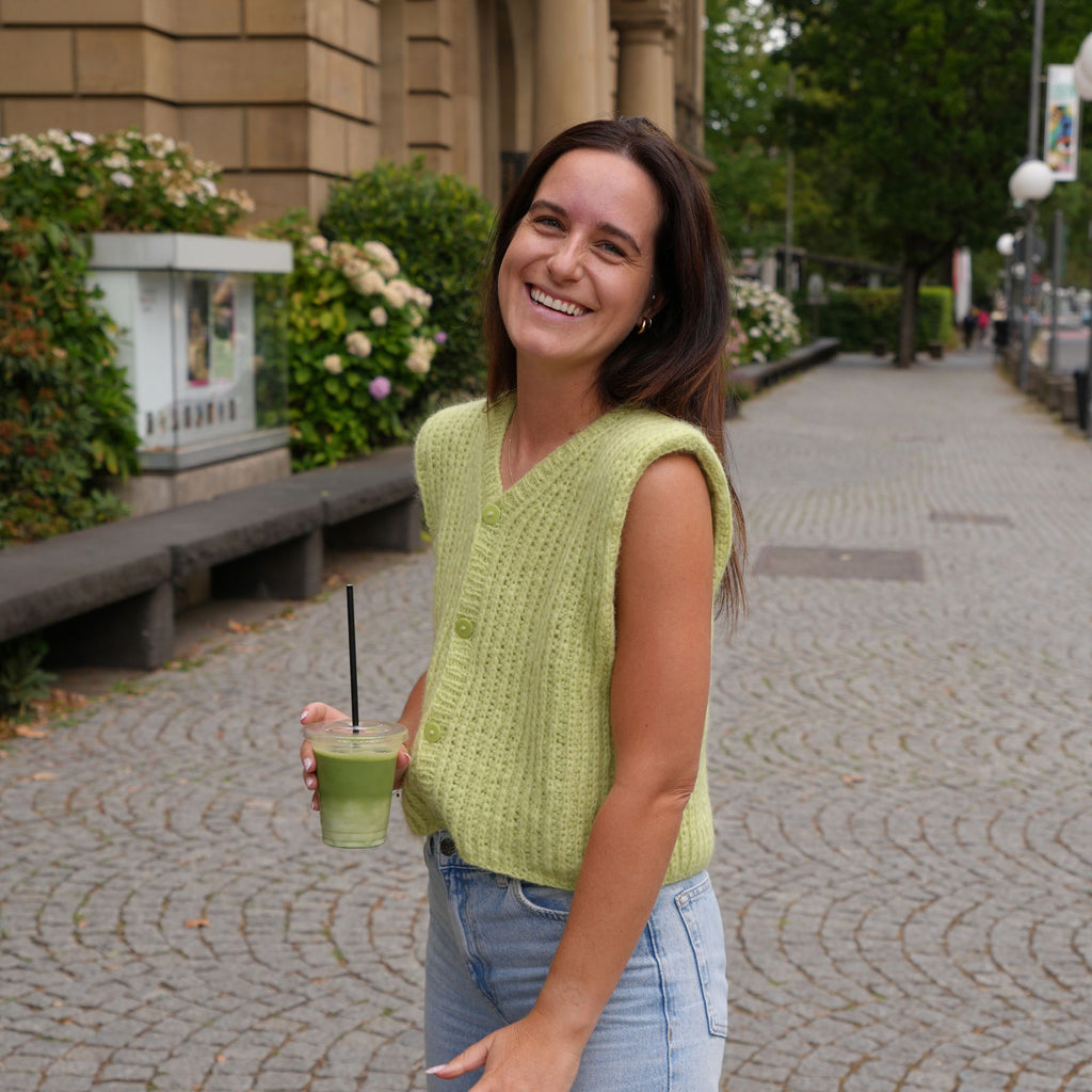 A woman smiling outdoors on a cobblestone street, wearing a sleeveless light green knit vest over a white top and light blue jeans, while holding a green drink with a black straw.