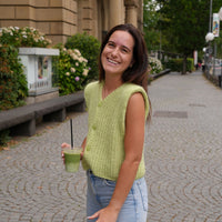 A woman smiling outdoors on a cobblestone street, wearing a sleeveless light green knit vest over a white top and light blue jeans, while holding a green drink with a black straw.