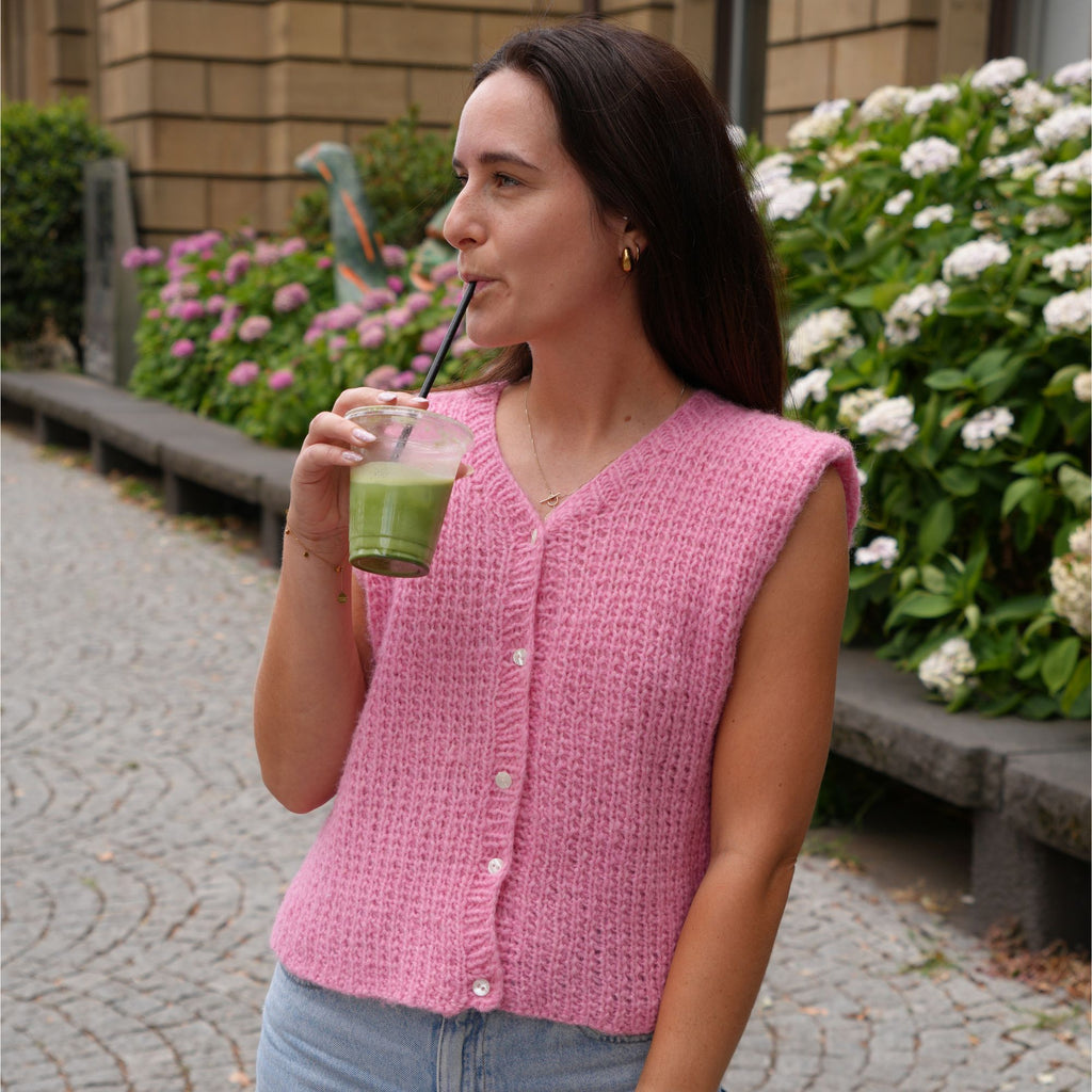 A woman wearing a pink sleeveless knit cardigan vest with white buttons, standing outdoors on a cobblestone path and drinking from a green drink.