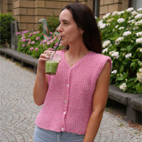 A woman wearing a pink sleeveless knit cardigan vest with white buttons, standing outdoors on a cobblestone path and drinking from a green drink.