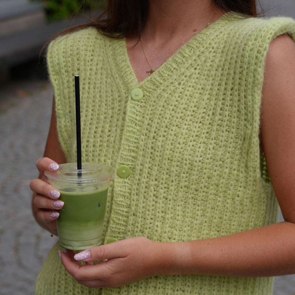 A person wearing a sleeveless light green knit vest and holding an iced matcha drink in a clear cup with a black straw.