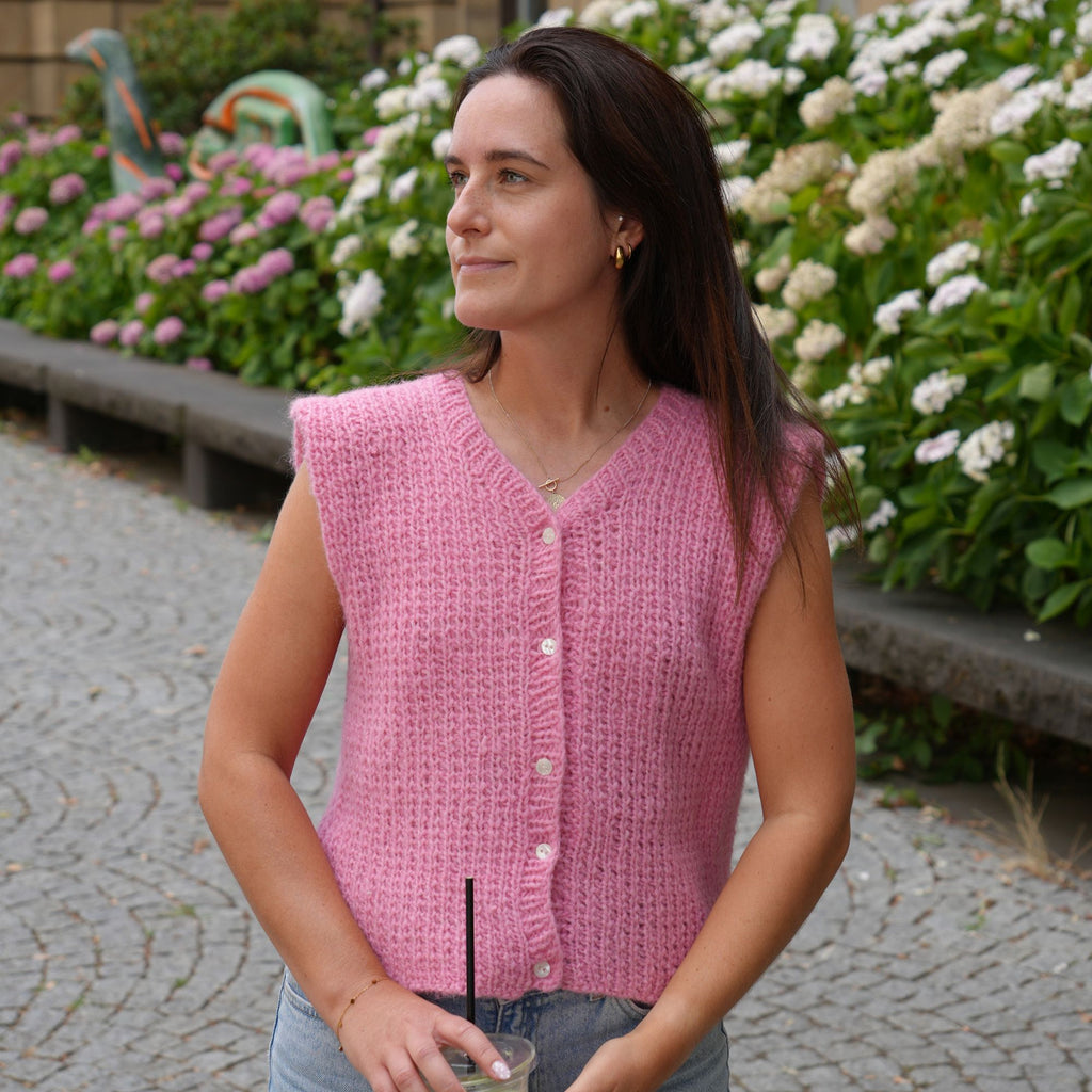 A woman wearing a pink sleeveless knit vest with white buttons, standing outdoors in front of a flower bed.