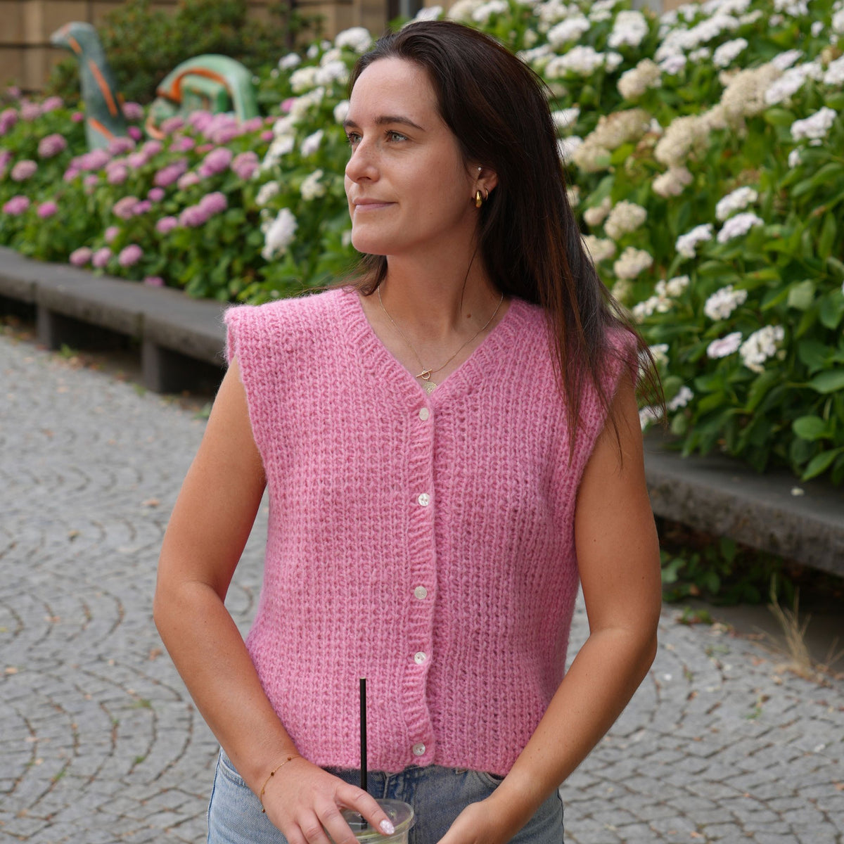 A woman wearing a pink sleeveless knit vest with white buttons, standing outdoors in front of a flower bed.