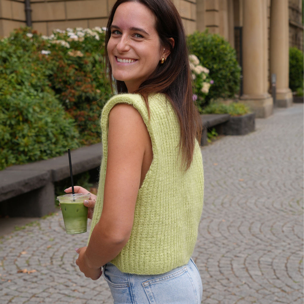 A woman wearing a sleeveless, textured light green knit vest and light blue jeans stands on a cobblestone path holding a green drink, looking back over her shoulder.